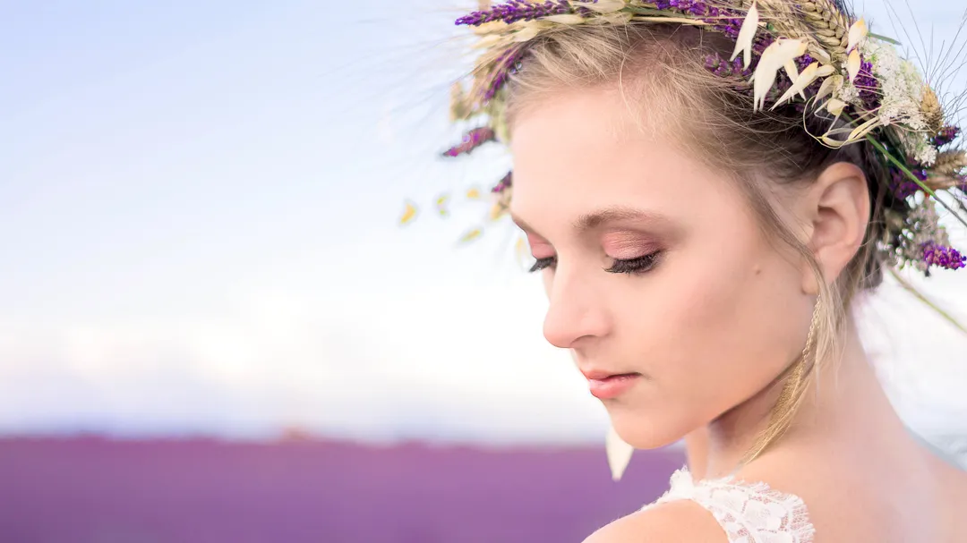 Femme aux cheveux blonds, le visage penché, portant une couronne de fleurs sauvages violettes sur un paysage montagneux violet.