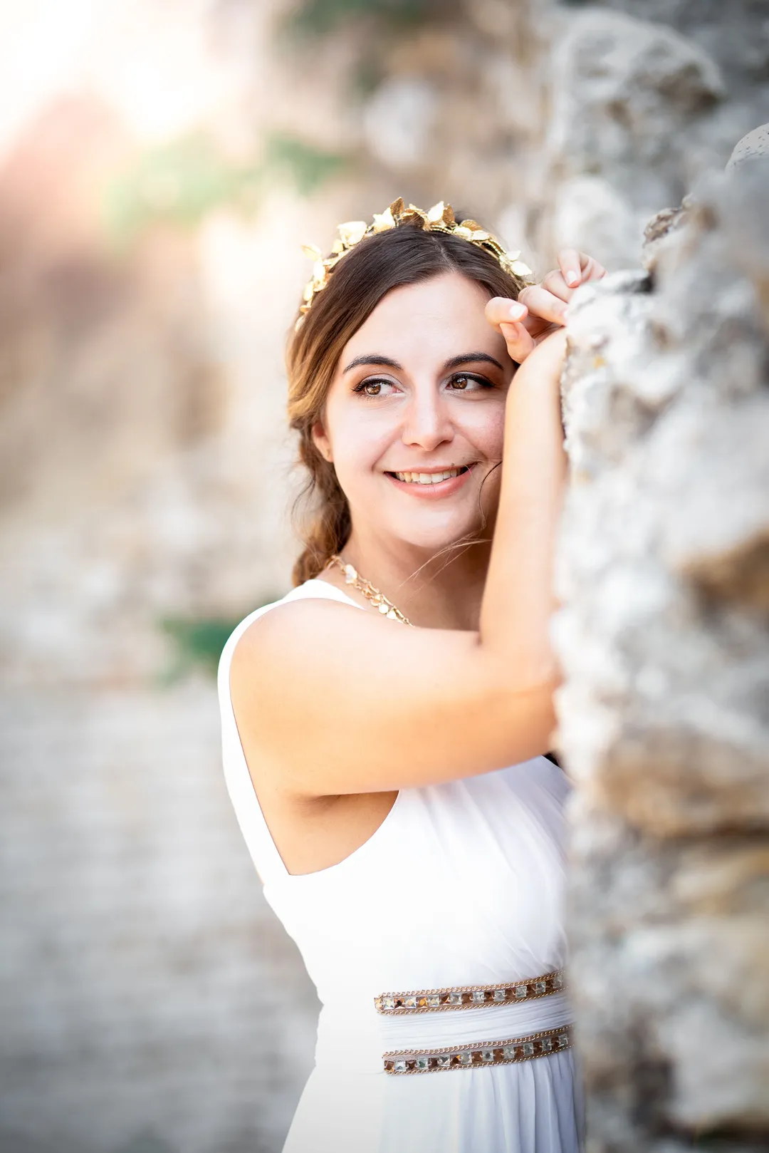 Jeune femme vêtue d'une longue robe blanche, se tenant contre un mur de pierre calcaire, avec une expression souriante et un diadème or sur la tête.