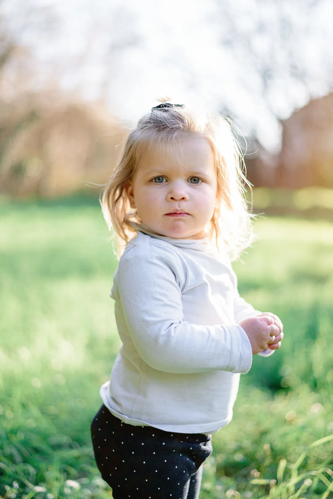 Une petite fille, vêtue d'un pull en laine blanche et de pantalons noirs et blancs, se tient debout dans un champ vert avec des arbres en arrière-plan. Elle regarde le spectateur avec un air sérieux.