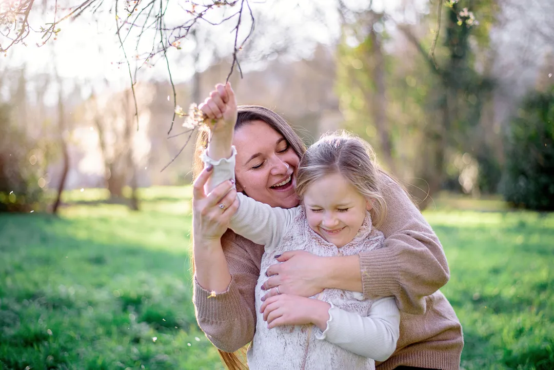 Mère et fille se câlinent dans un jardin printanier, entourées de fleurs et de feuilles éparpillées.