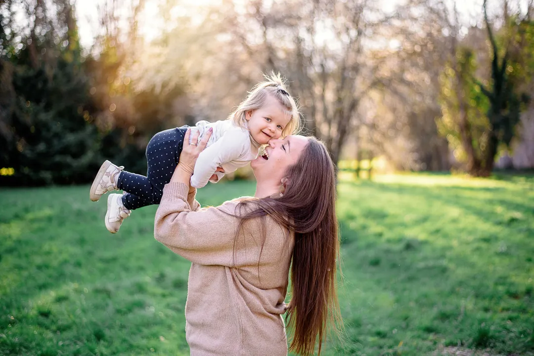 Une maman souriante soulève sa petite fille en l'air dans un parc verdoyant au printemps.