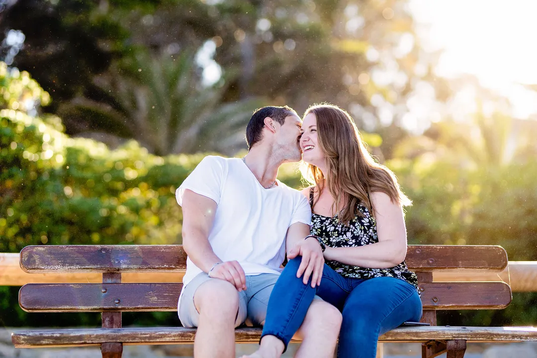 Couple souriant assis sur un banc en bois dans un jardin verdoyant, baigné de lumière dorée.
