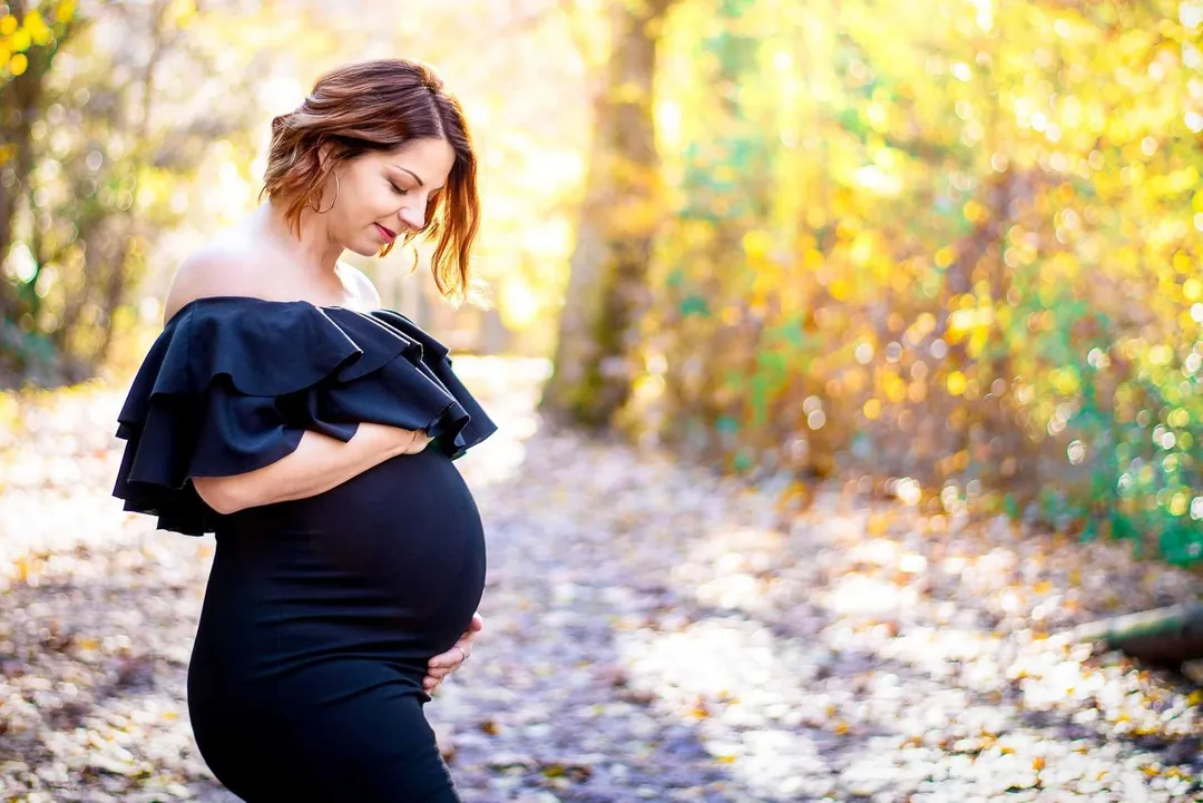 Femme enceinte se tenant le ventre, dans un sentier recouvert de feuilles mortes automnales, lumière filtrée.