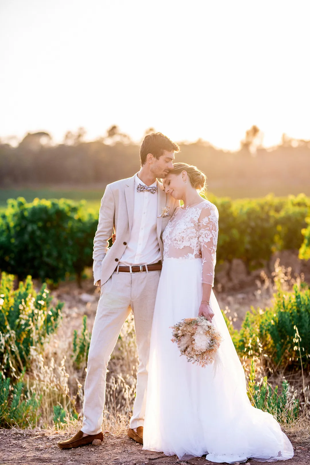 Mariés dans un décor champêtre, le couple se tient debout dans un terrain vague planté de vignes. Le mari porte un costume beige et le mariée une robe blanche. Ils serraient la main et sont proches l'un de l'autre
