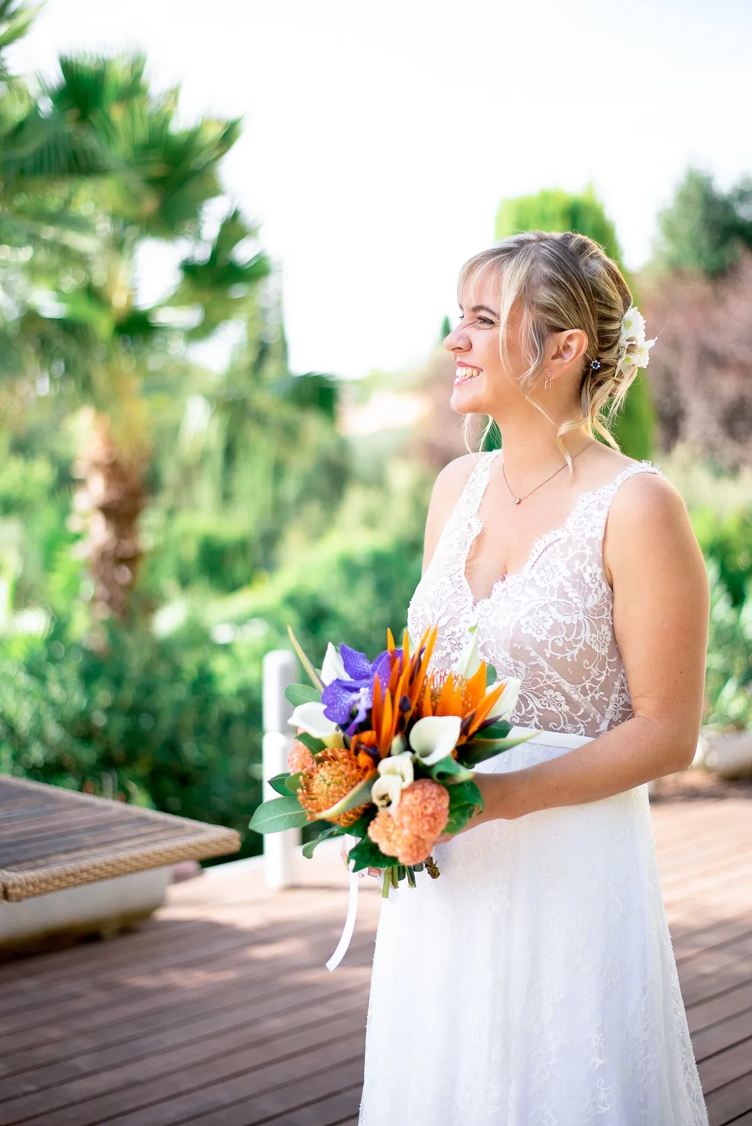 Mariée souriante tenant son bouquet de fleurs exotiques sur une terrasse en bois ombragée par des palmiers.