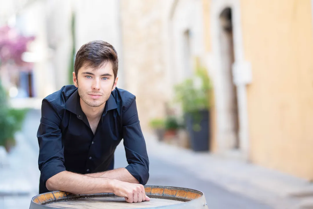 Un jeune homme aux cheveux bruns, vêtu d'une chemise noire, se tient dans une ruelle pittoresque avec des murs blanchis à la chaux et des arbres en arrière-plan.