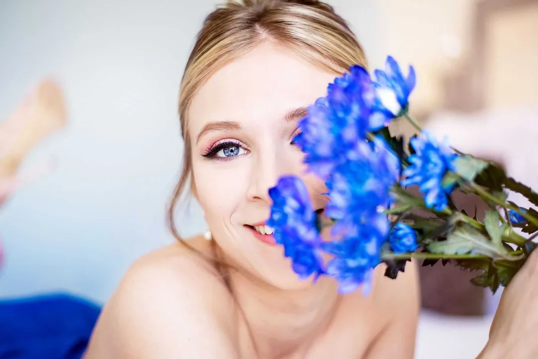 Jeune femme blonde souriante tenant un bouquet de myosotis bleus, arrière-plan blanc et neutre.