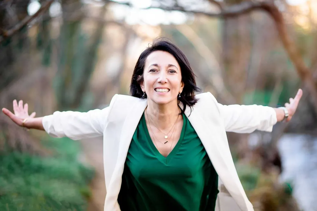 Femme souriante, les bras ouverts, en tailleur blanc et chemise verte, dans un environnement boisé et verdoyant.