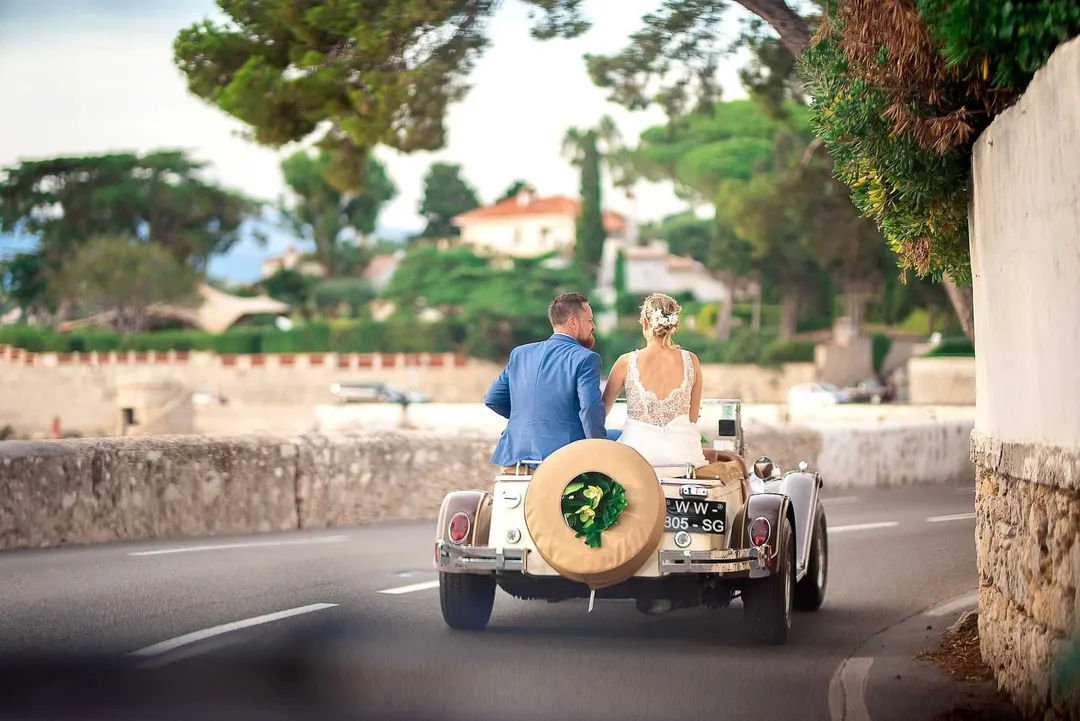 Mariage : la mariée et le marié sur une ancienne voiture vintage, avec un grand volant en rotin, sur une route bordée d'arbres et d'un remblai, avec une maison en arrière-plan.