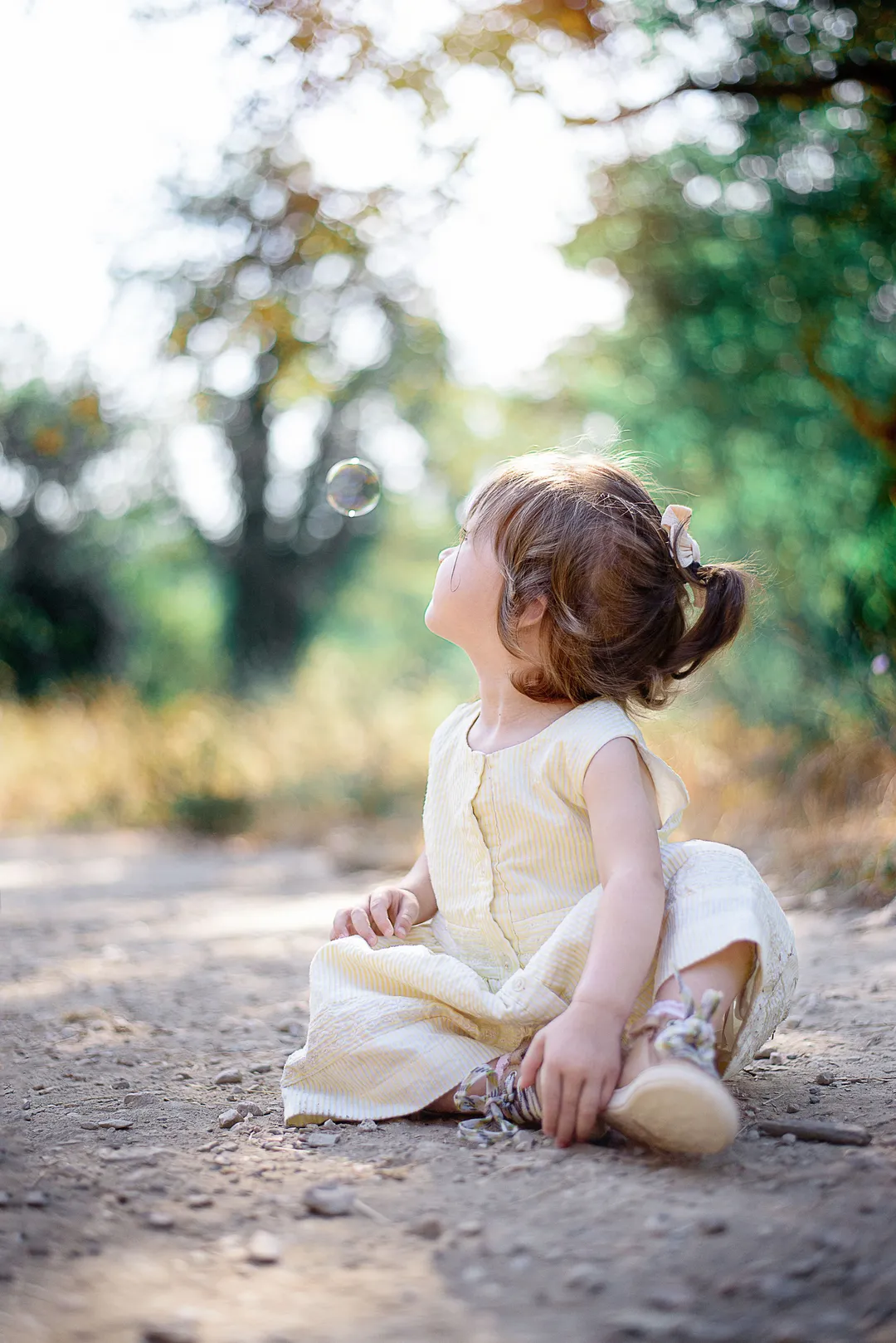 Une jeune fille avec des cheveux bruns attachés en chignon, vêtue d'une robe jaune clair, assise sur un chemin de terre sous un arbre feuillu.