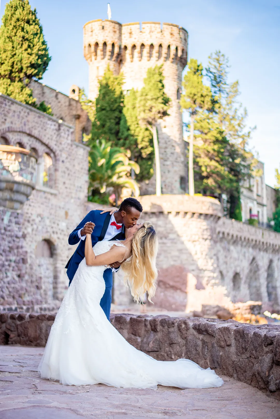 Mariée et marié seembrassent dans un décor de château, avec des marches de pierre et des arbres en arrière-plan.