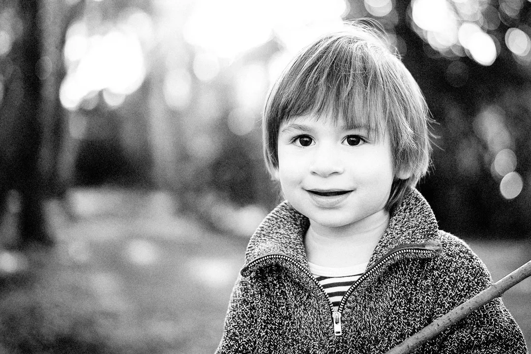 Enfant souriant, portant un gilet en laine et un bâton, dans un environnement boisé flou.
