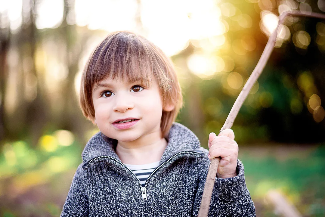 Enfant avec des cheveux bruns, portant un pull gris et un pull rayé noir et blanc, tenant un bâton dans une main, un sourire malicieux.