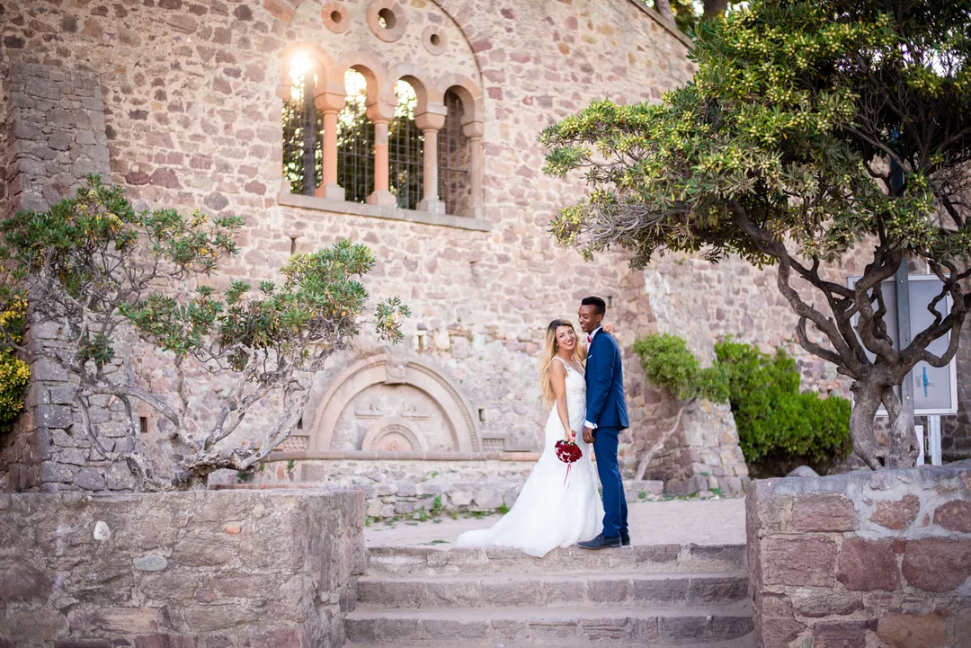 Mariée et marié se tenant la main sur les escaliers devant une façade en pierre avec des arcatures.