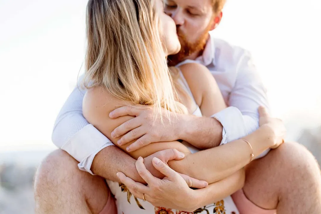 Couple se tenant tendrement sur le sable, une femme s'enroule autour de l'homme, leurs visages proches dans un moment de tendresse.