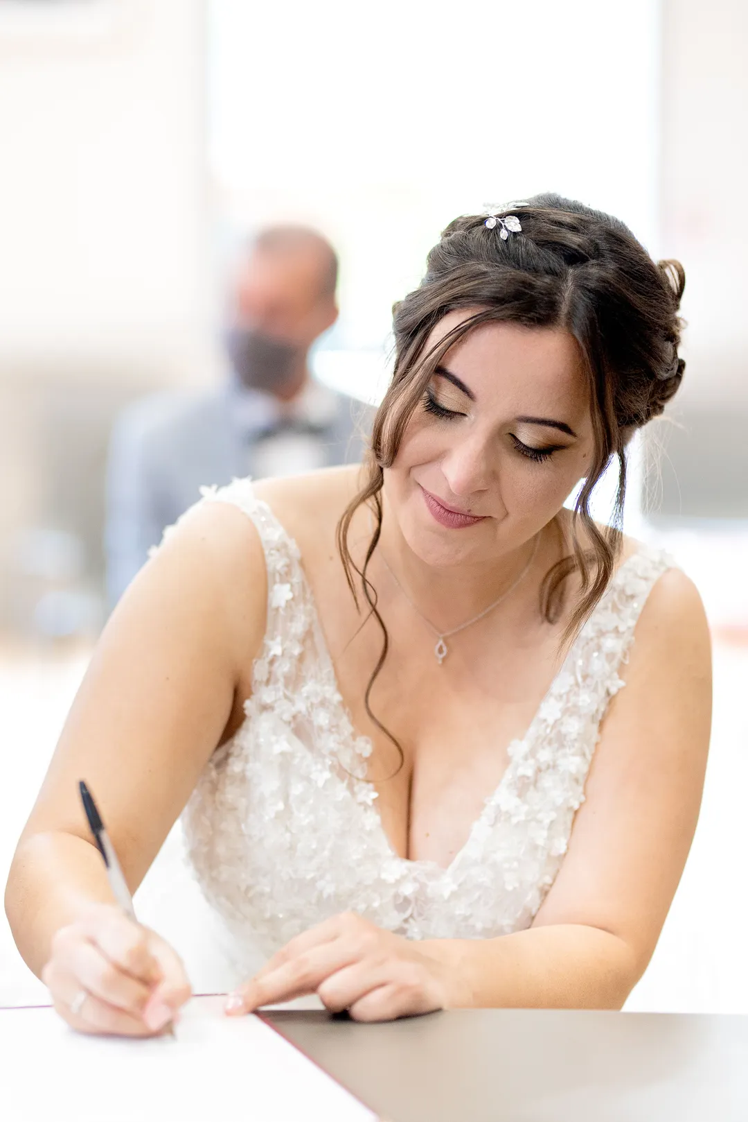Une jeune mariée, vêtue d'une robe en dentelle blanche, signe un registre à l'aide d'un stylo. Un homme est visible en arrière-plan.