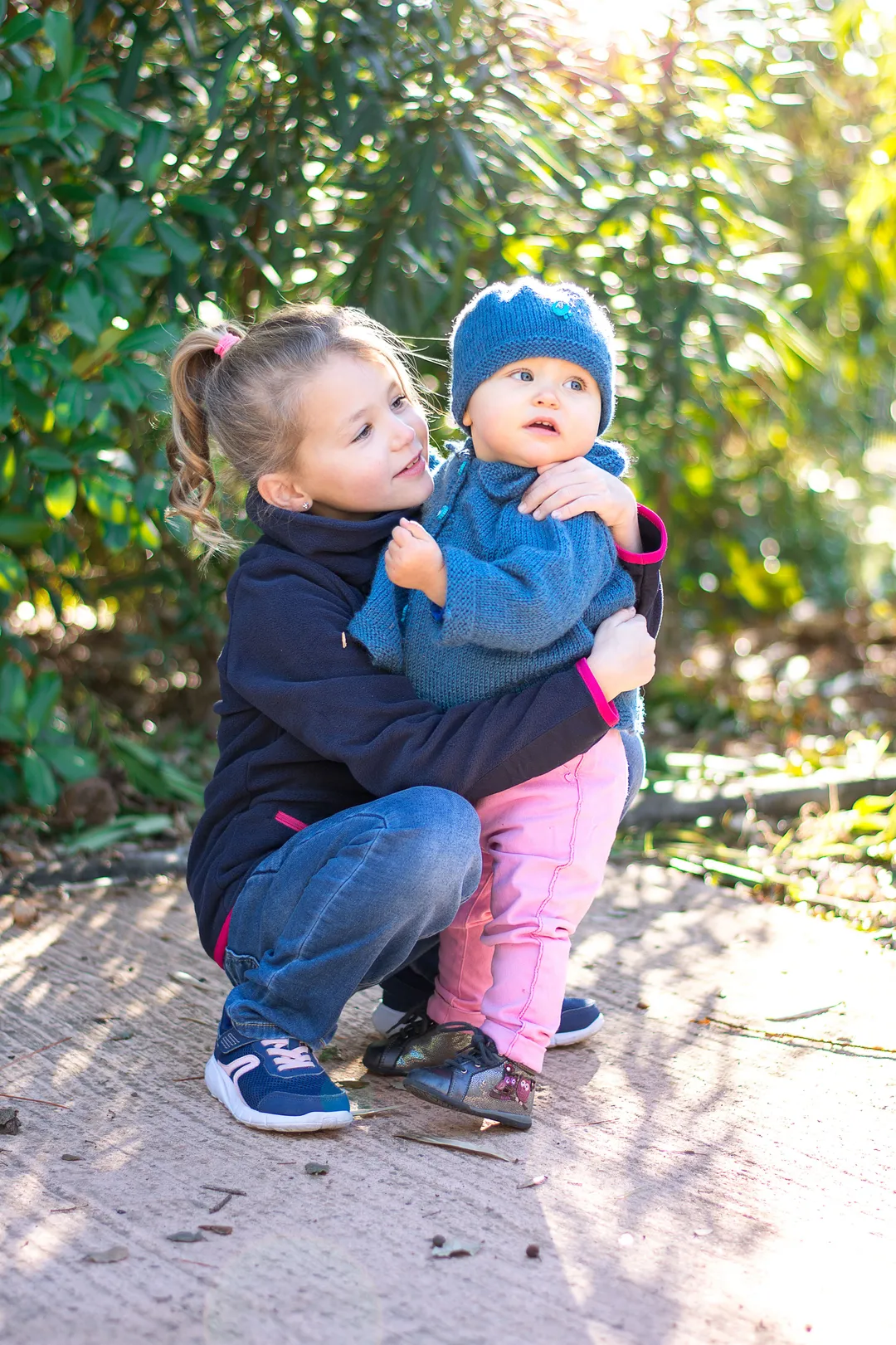 Deux jeunes filles, une grande et une petite, se tiennent dans un parc verdoyant. La plus petite est dans les bras de sa grande sœur.