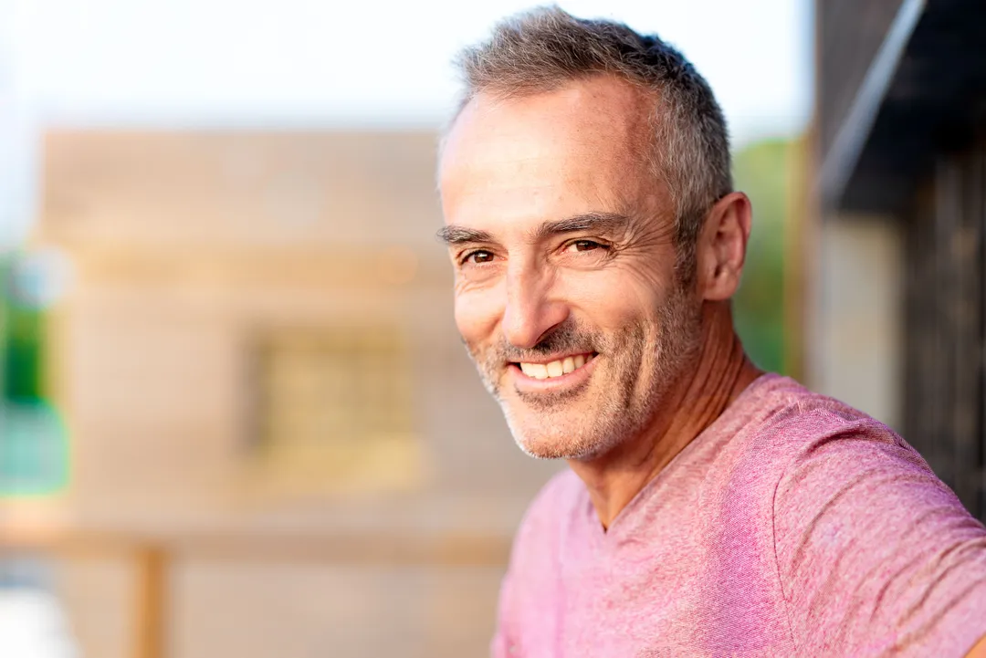 Homme souriant avec une barbe, portant un t-shirt rose, devant un mur en bois.