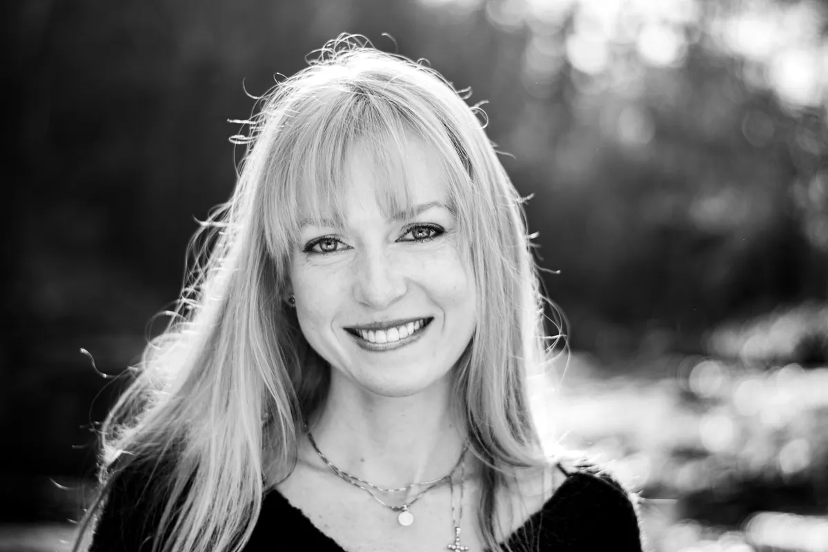 Portrait en noir et blanc d'une jeune femme aux cheveux blonds longs et ondulés, souriante, en extérieur avec des arbres en arrière-plan.