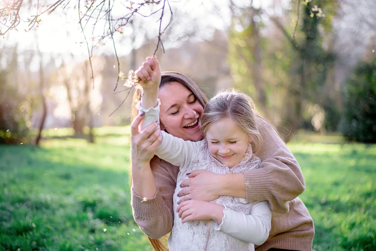 Mère et fille se câlinent dans un jardin printanier, entourées de fleurs et de feuilles éparpillées.
