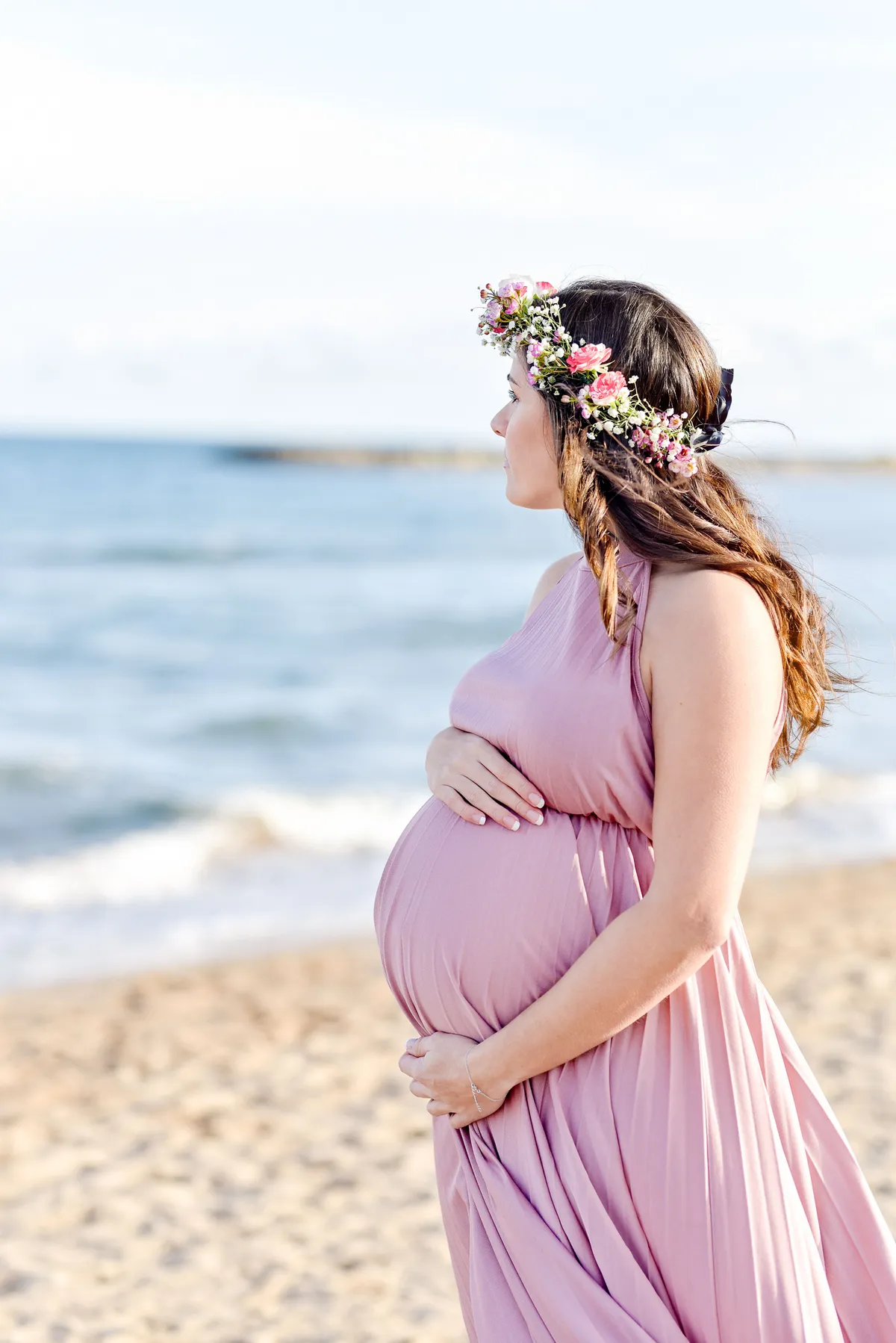 Femme dans un long voile rose sur une plage, regardant vers l'horizon avec des vagues bleues au loin.