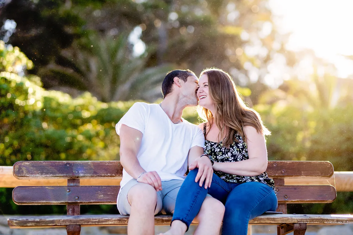 Couple souriant assis sur un banc en bois dans un jardin verdoyant, baigné de lumière dorée.