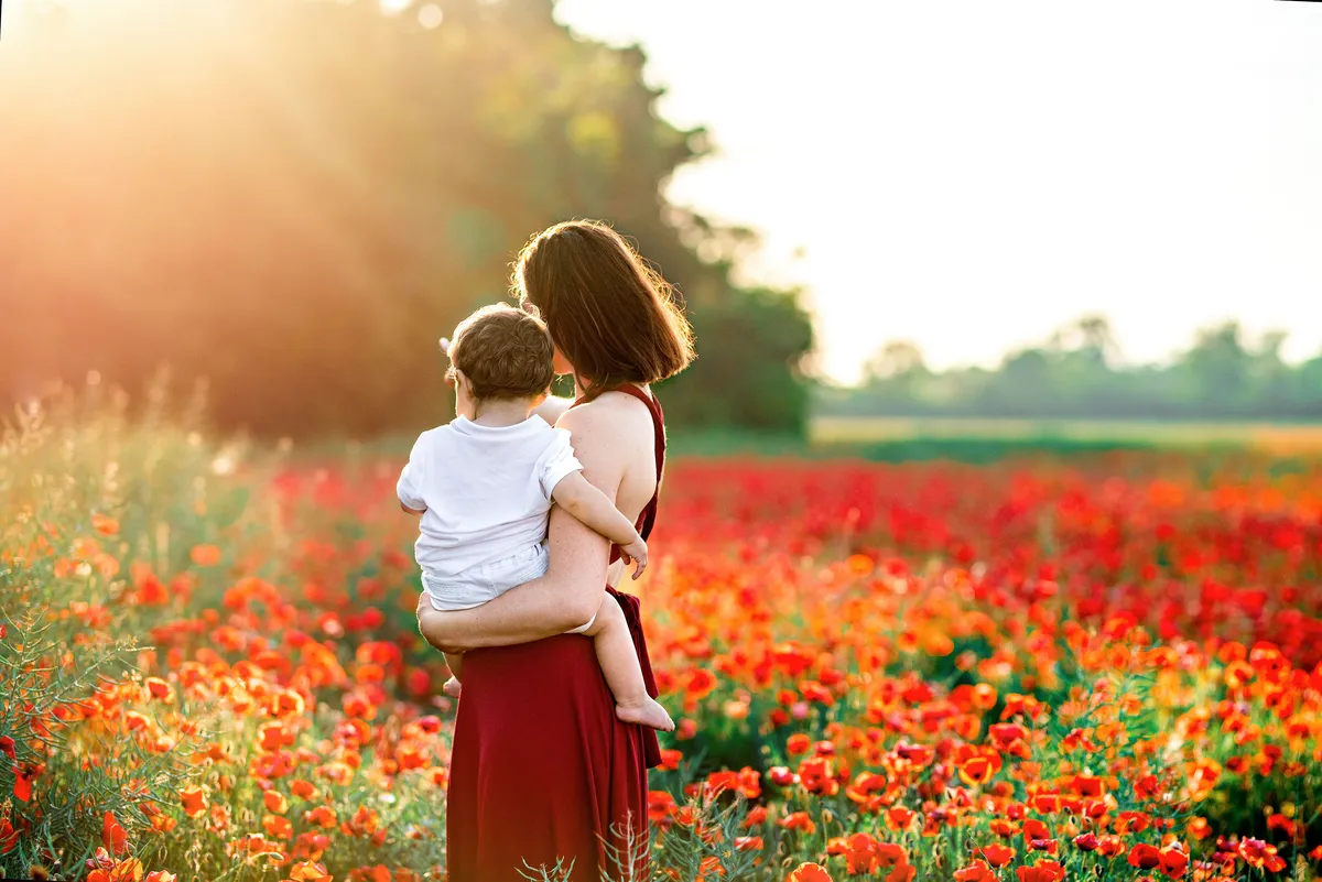 Une mère porte son enfant dans ses bras, debout dans un champ de coquelicots rouges en fleur au coucher du soleil.