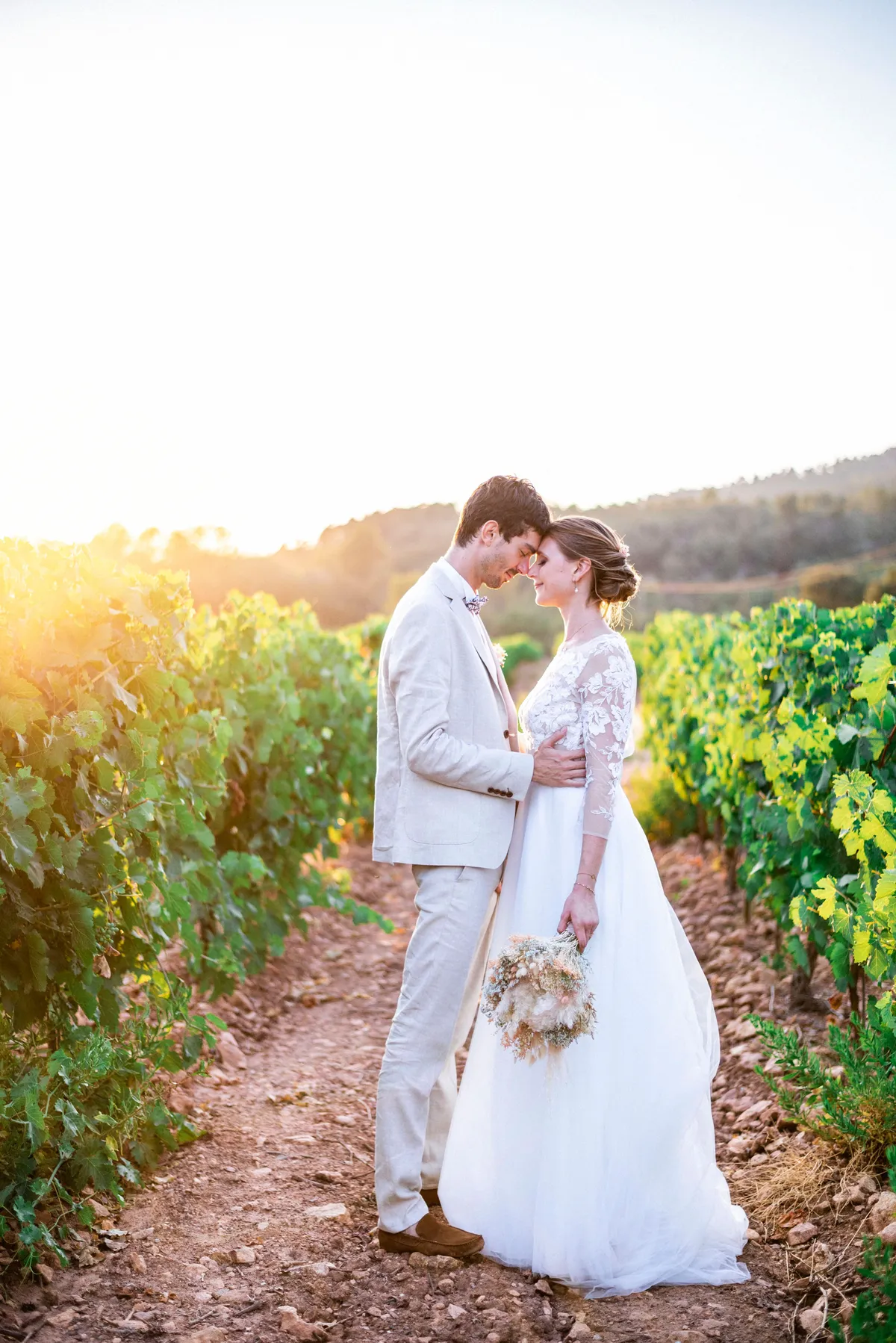 Jeune couple marié se tenant et se regardant dans une vignoble ensoleillé. La mariée porte une robe de mariée longue blanche et le marié un costume beige.