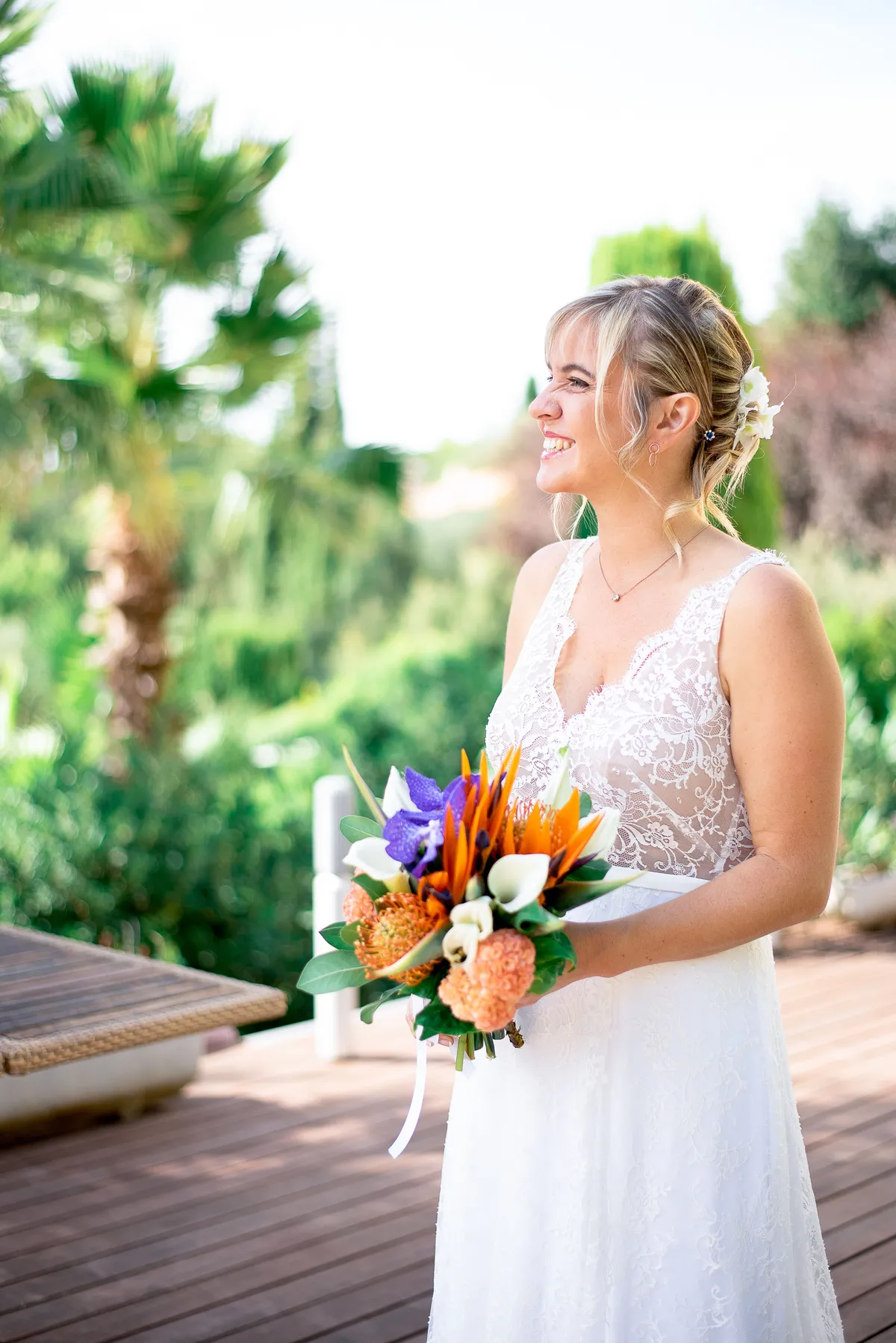 Mariée souriante tenant son bouquet de fleurs exotiques sur une terrasse en bois ombragée par des palmiers.