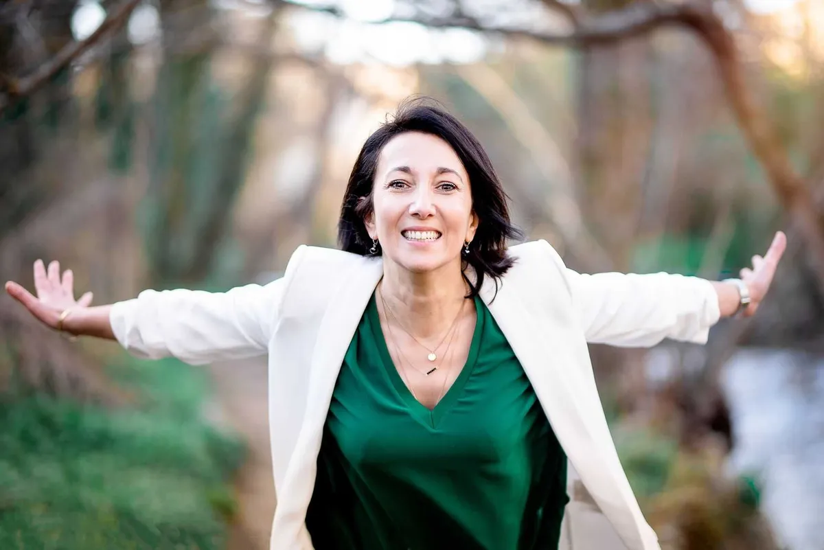 Femme souriante, les bras ouverts, en tailleur blanc et chemise verte, dans un environnement boisé et verdoyant.