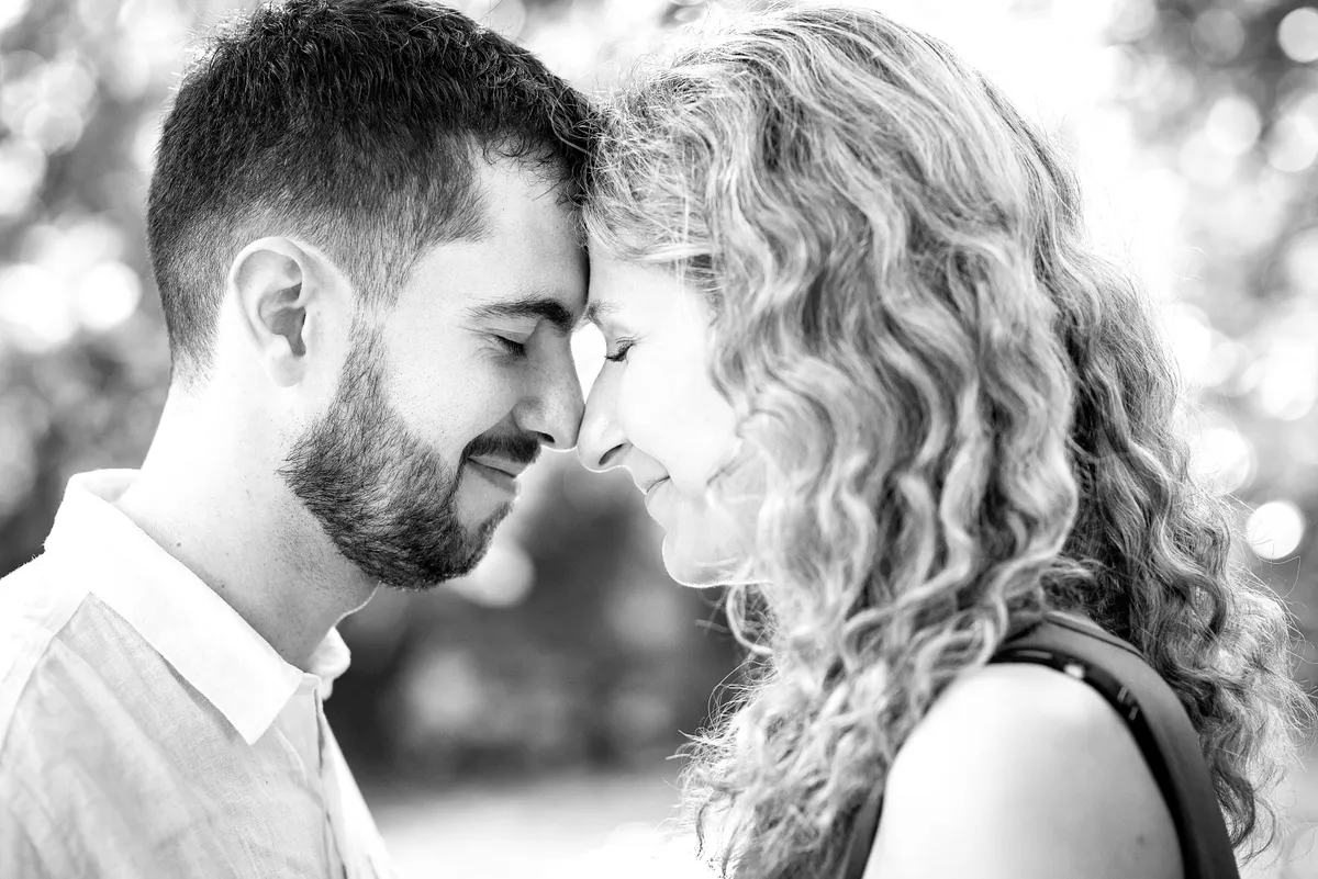 Couple se regardant tendrement, leurs visages se touchant. Arbres verdâtres en arrière-plan.