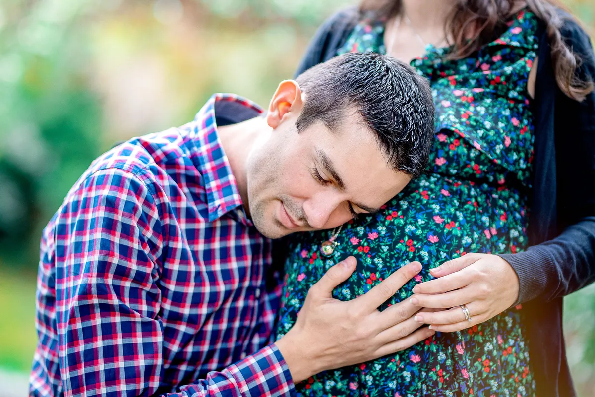 Un homme enceinte, le regard rêveur, pose sa main sur le ventre de sa femme. Il porte une chemise à carreaux rouge et noir et un pull noir.