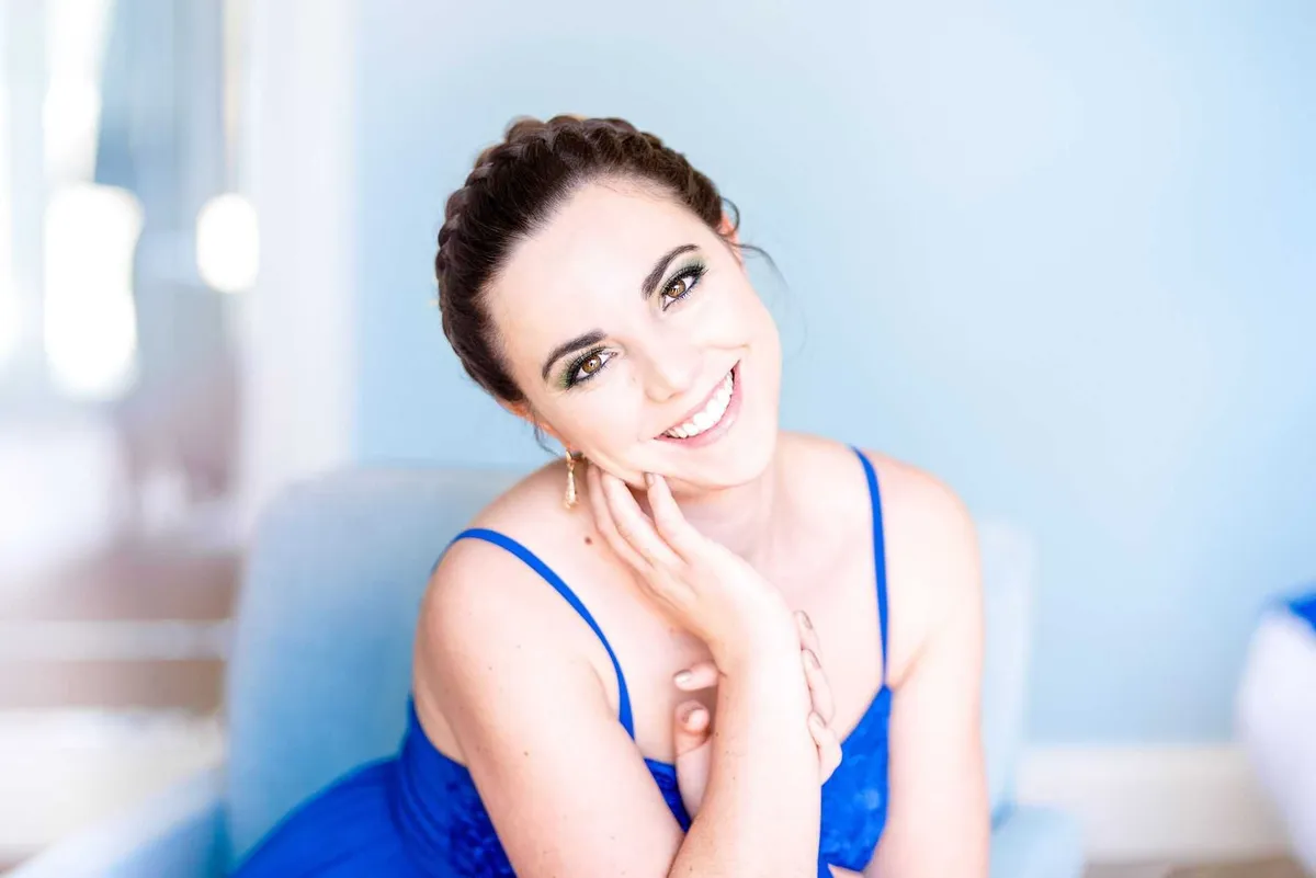 Jeune femme souriante, vêtue d'une robe bleu marine scintillante, elle se tient assise et pose sa main sur son visage.