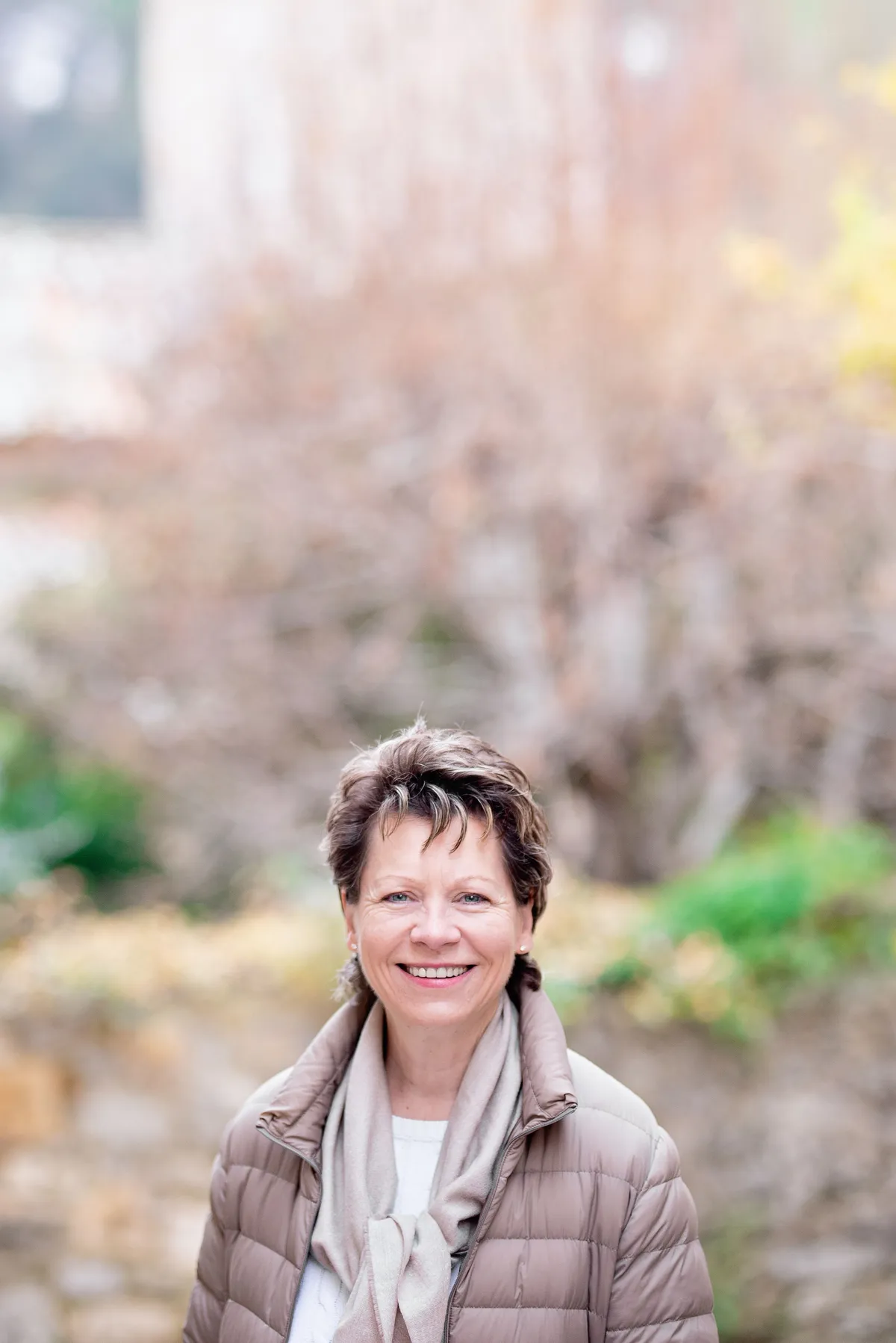 Femme d'âge mûr souriante, portant un manteau matelassé beige et un foulard gris, se tient devant un arrière-plan de végétation automnale.