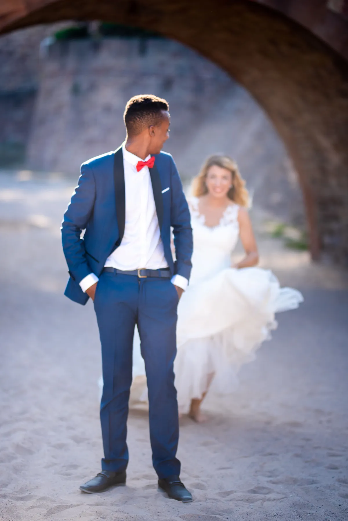 Un photographe en costume bleu marine regarde une mariée en robe blanche éthérée se détacher dans un espace urbain.