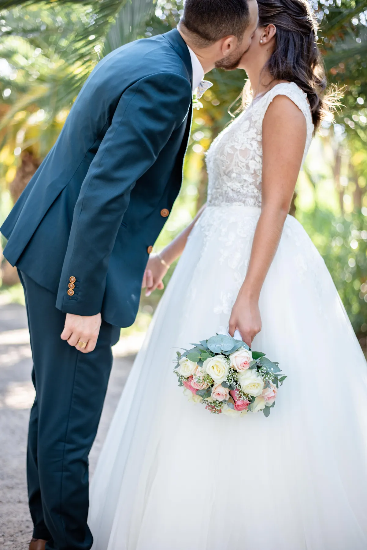 Homme en costume sombre, tenant un bouquet de roses et de pivoines, se penche pour embrasser la mariée vêtue d'une robe blanche.