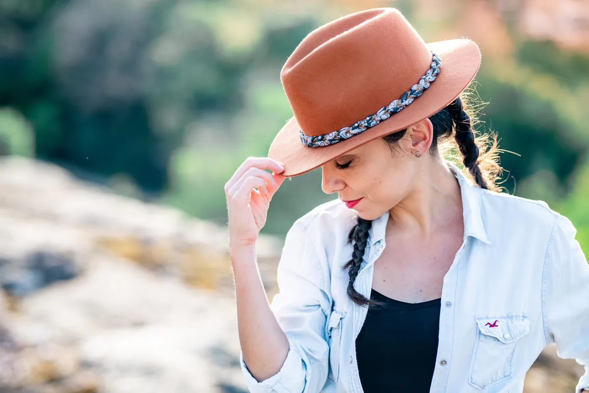 Femme avec des cheveux tressés, portant un jean déimbré et un chapeau en feutre brun, debout dans un paysage montagneux.