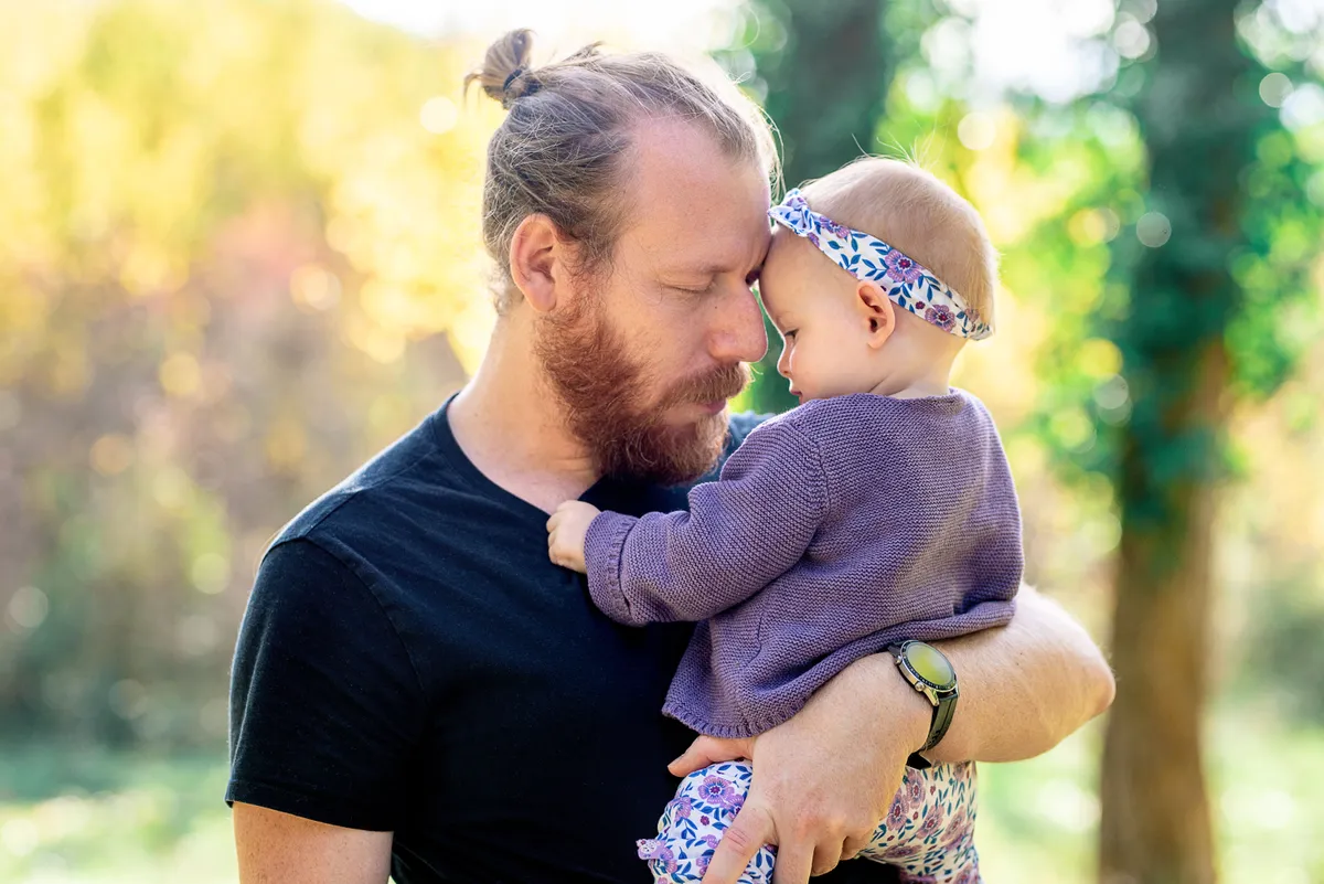 Un père tient sa fille dans ses bras, les visages proches, dans un environnement boisé en lumière.