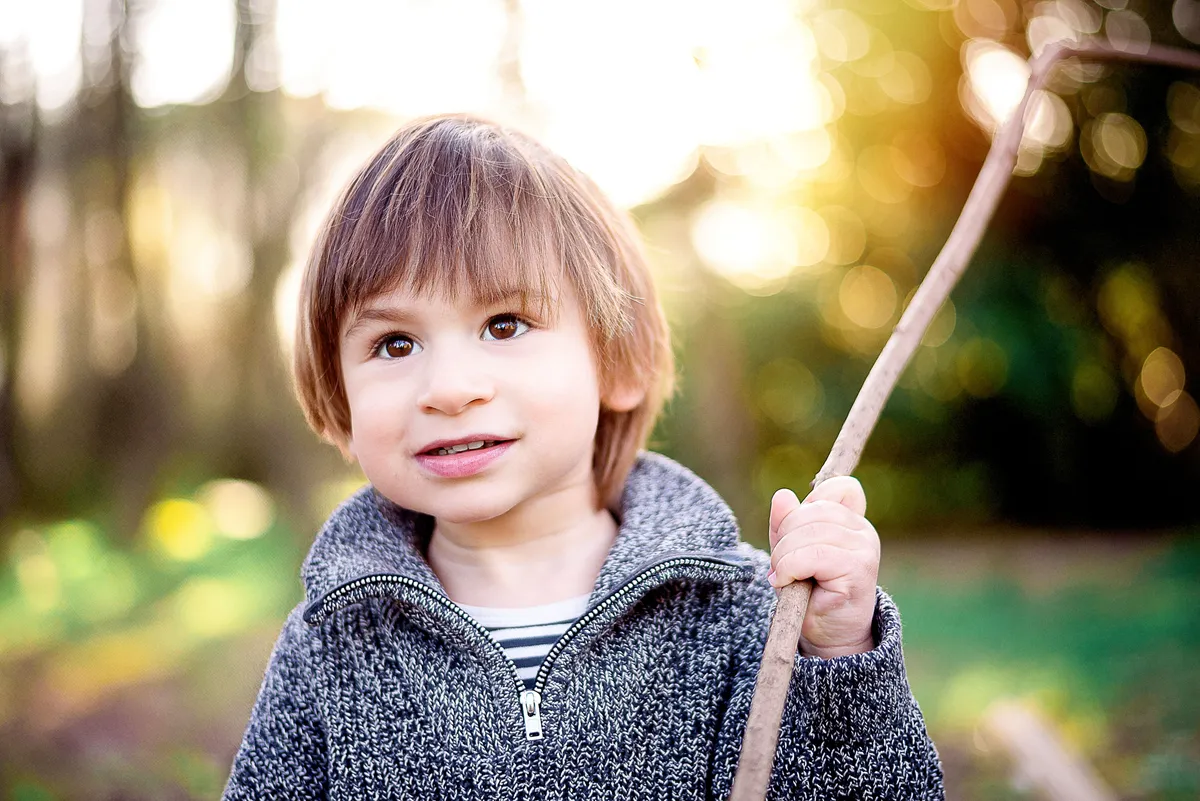 Enfant avec des cheveux bruns, portant un pull gris et un pull rayé noir et blanc, tenant un bâton dans une main, un sourire malicieux.