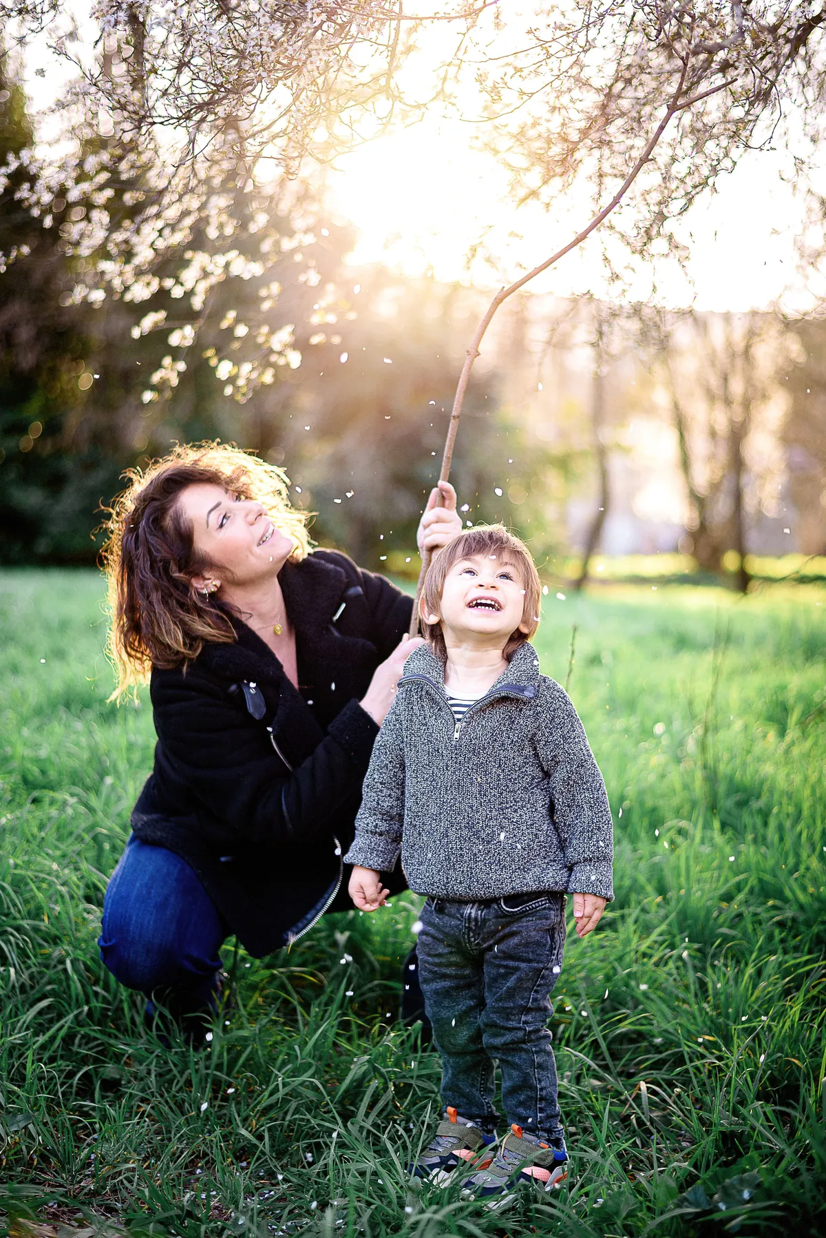 Un jeune garçon, souriant et excité, souffle des pétales de fleurs blanches dans un champ verdoyant au coucher du soleil.