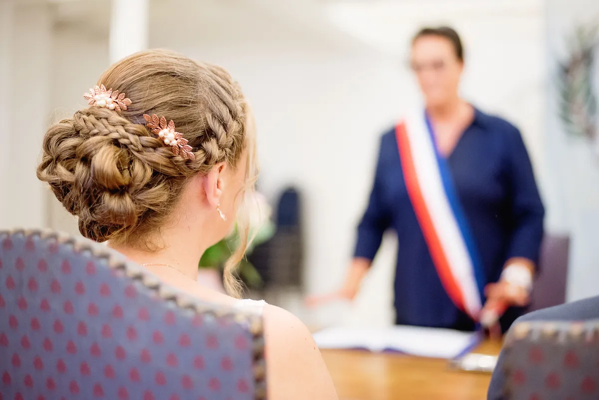 Une jeune femme aux cheveux tressés, assise dos au spectateur, travaille à une table avec un homme en costume bleu et un drapeau tricolore.