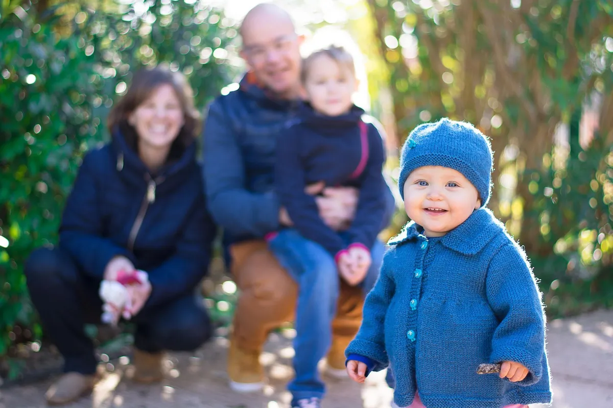 Un enfant en manteau bleu, avec un petit bâton, se tient devant une famille assise dans un jardin feuillu.