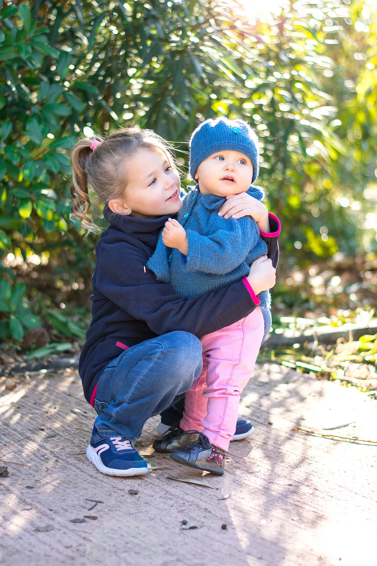 Deux jeunes filles, une grande et une petite, se tiennent dans un parc verdoyant. La plus petite est dans les bras de sa grande sœur.