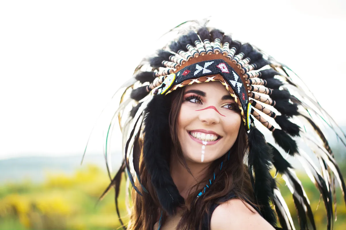 Femme portant un grand plumeau de chef en plumes noires et blanches, avec du maquillage tribal sur le visage, paysage de fond verdoyant